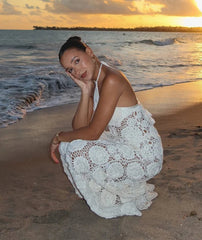 Woman in a white lace dress sitting on a beach at sunset.