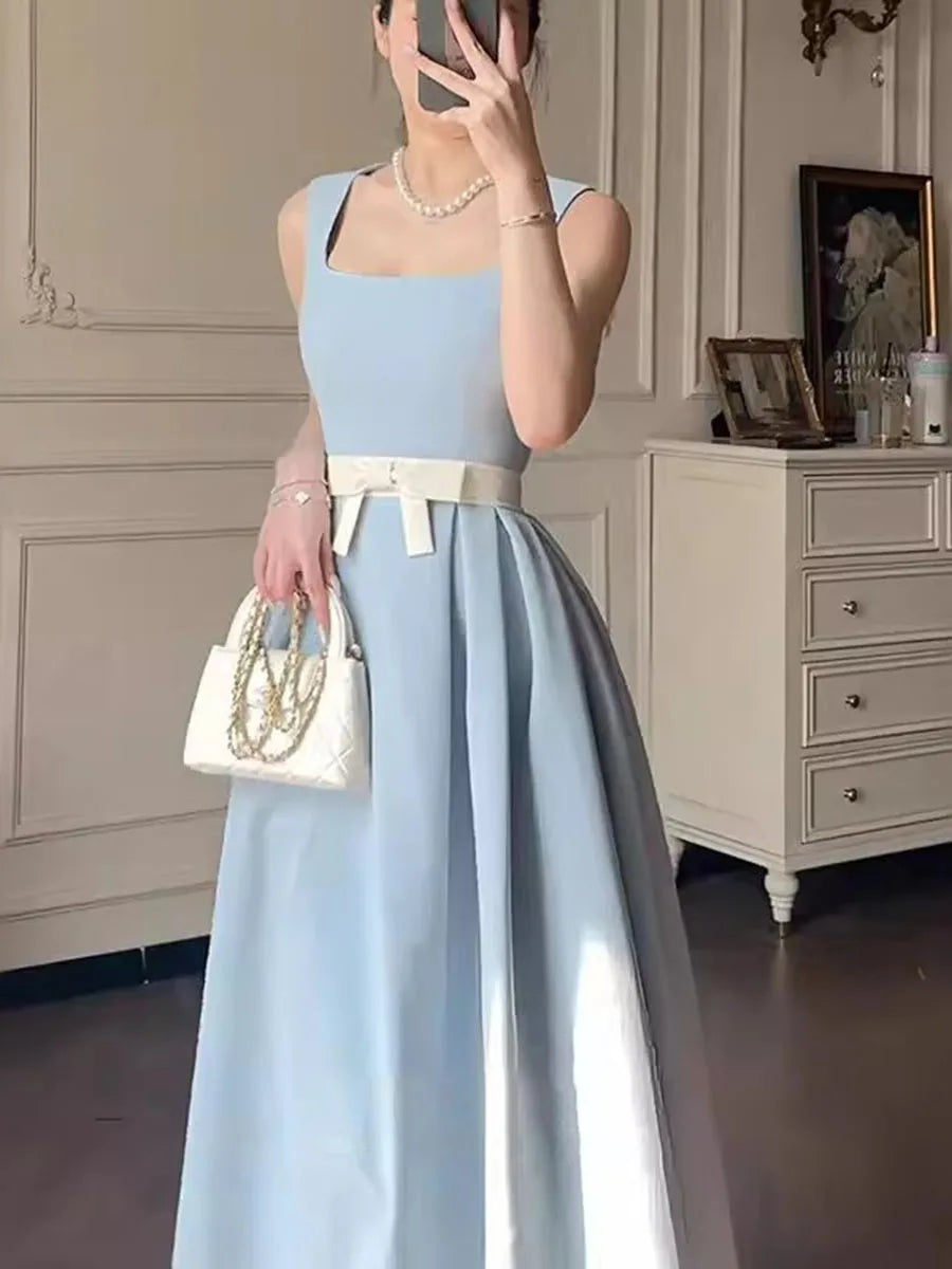 Woman in a light blue dress with a white belt, holding a white handbag, standing in a room.