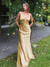 Woman in a gold evening gown standing outdoors with greenery in the background