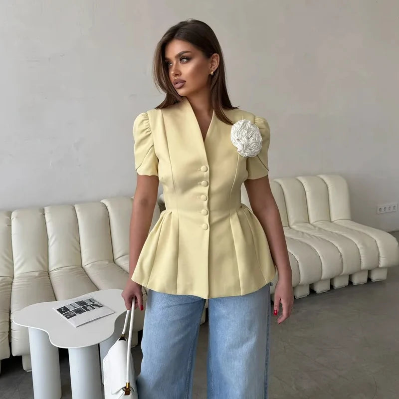 Woman wearing a light yellow peplum jacket with a white flower accessory, standing in a room with beige sofas.