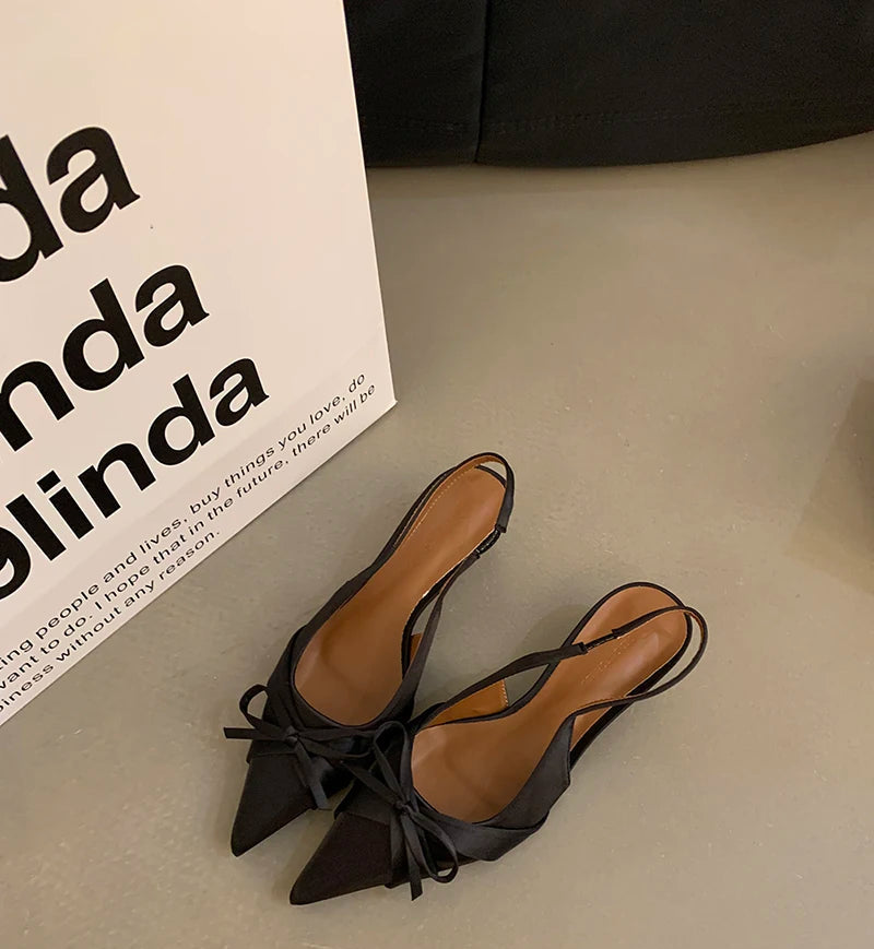 Black pointed-toe shoes with bow detail on a light gray surface, with a branded box in the background.