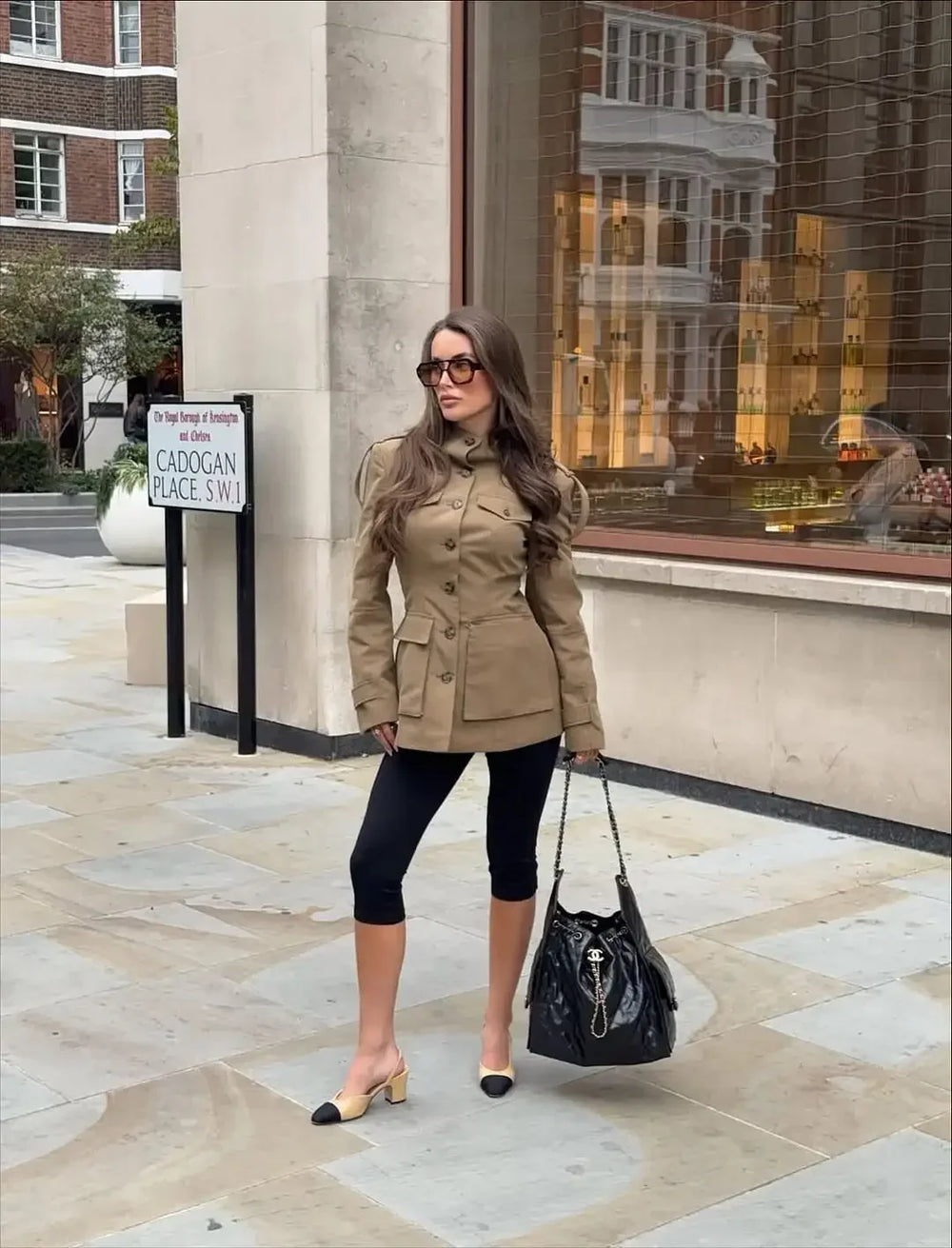 Woman in a beige coat and black pants standing on a stone pavement with a building in the background.