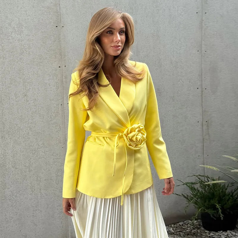 Woman wearing a yellow blazer with a floral detail against a gray wall.