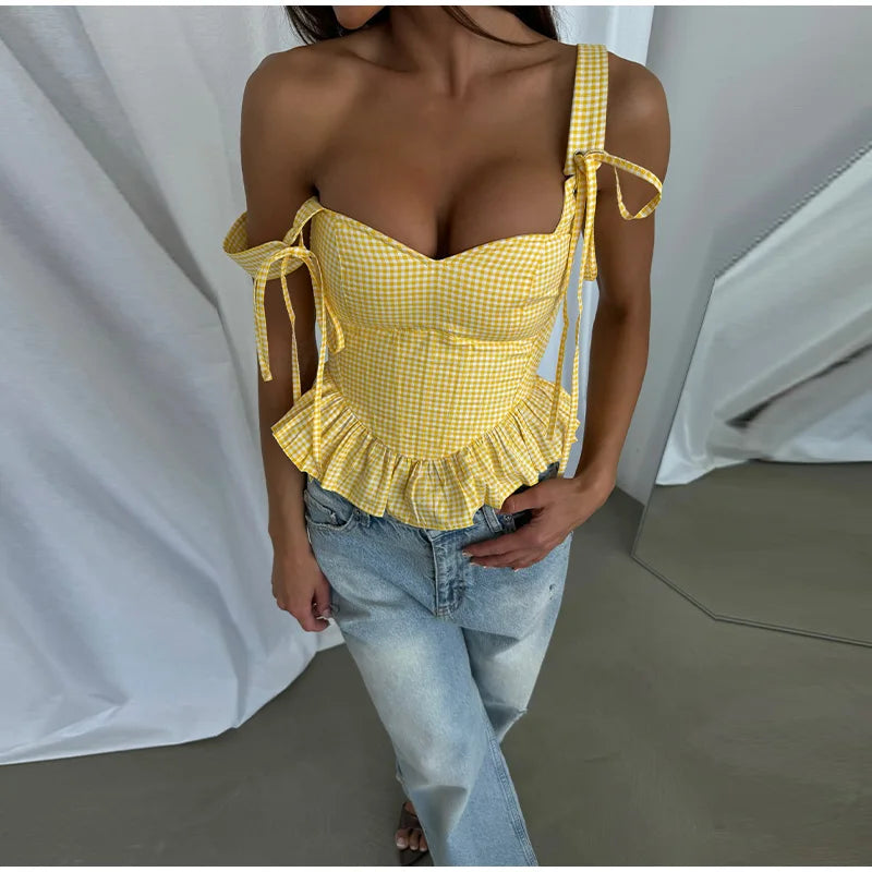 Person wearing a yellow checkered top with ruffled details and light blue jeans against a white curtain background.