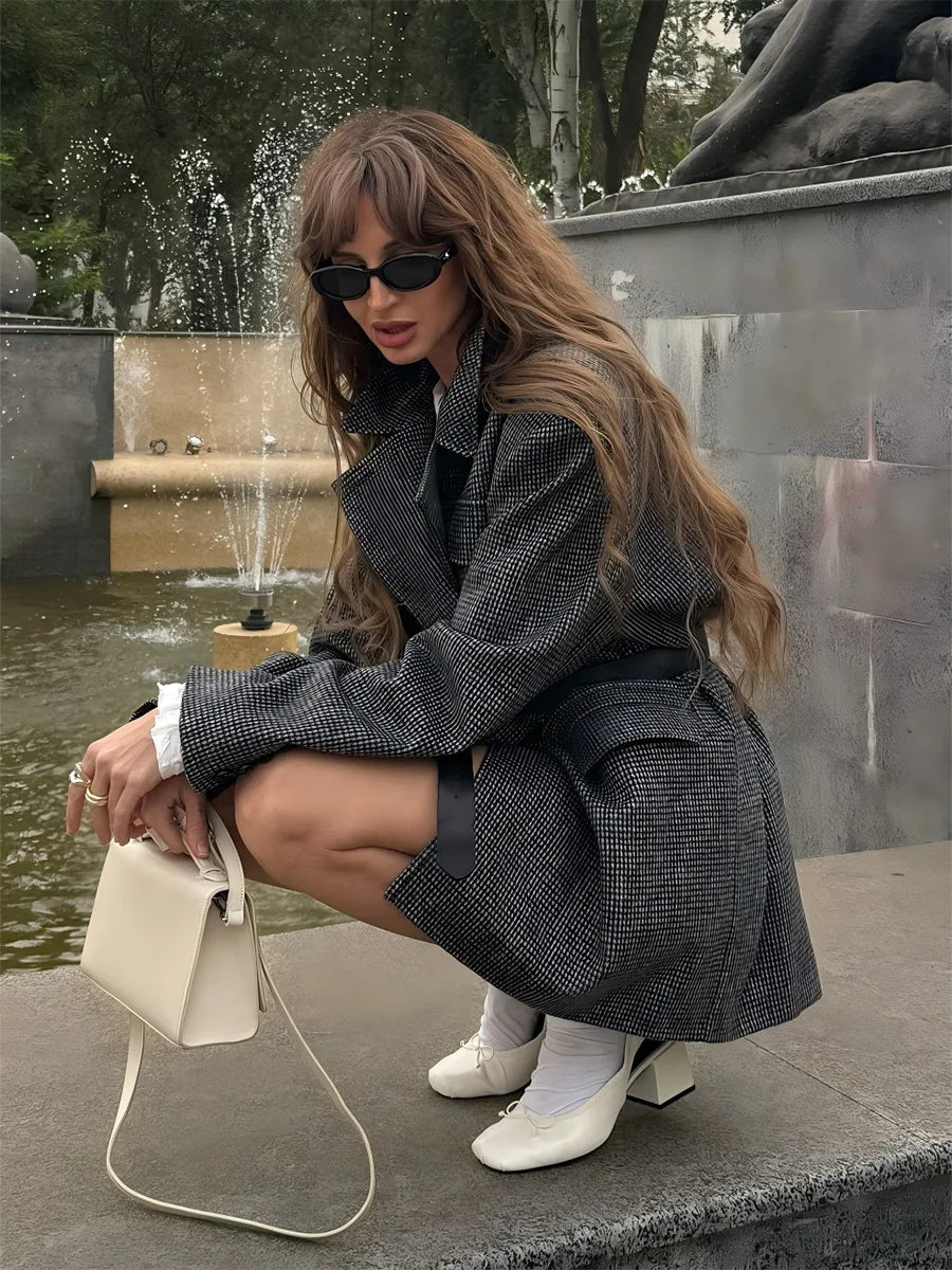 Woman in a gray coat and sunglasses sitting by a fountain