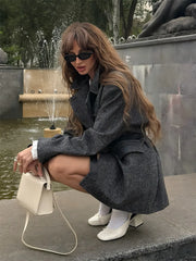 Woman in a gray coat and sunglasses sitting by a fountain