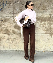 Person wearing a white blouse and brown pants against a textured wall.