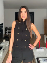 Woman wearing a black sleeveless jacket with gold buttons in a modern kitchen.