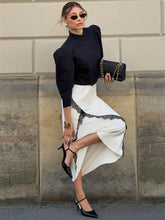 Woman in black top and white skirt with sunglasses and clutch against a stone wall.