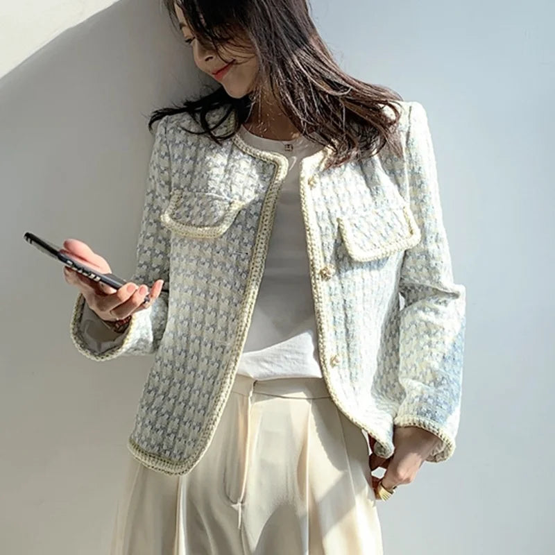 Woman wearing a light-colored textured jacket and beige pants, holding a phone against a plain background.