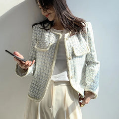 Woman wearing a light-colored textured jacket and beige pants, holding a phone against a plain background.