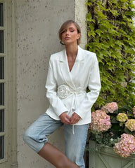 Woman wearing a white blazer with floral details and light blue jeans, standing against a textured wall with greenery.