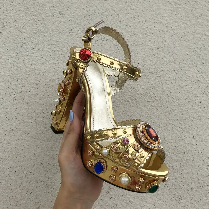 Gold high-heeled shoe with gemstone embellishments held by a hand against a neutral background