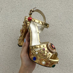 Gold high-heeled shoe with gemstone embellishments held by a hand against a neutral background