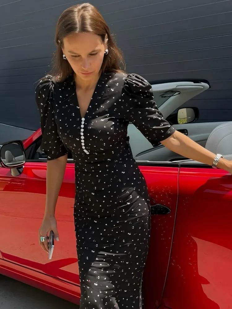 Woman in a black dress with white patterns standing next to a red car.