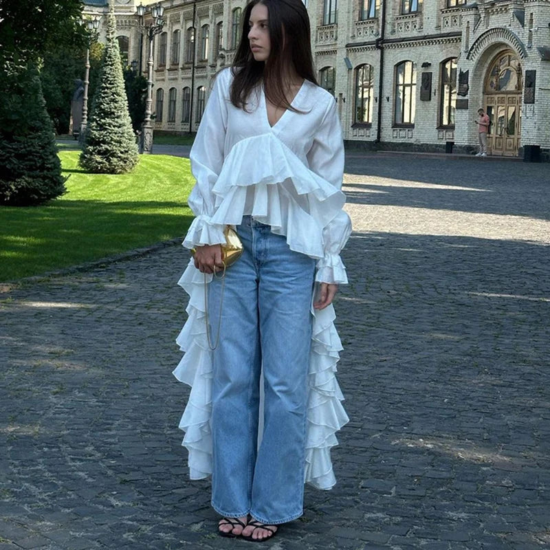 Woman wearing a white ruffled blouse and blue jeans standing on a cobblestone street with a building in the background.