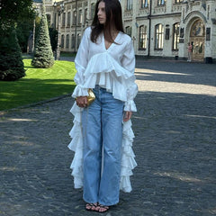 Woman wearing a white ruffled blouse and blue jeans standing on a cobblestone street with a building in the background.