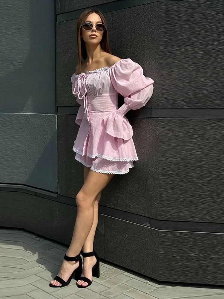 Woman in a pink off-shoulder dress standing against a dark wall.