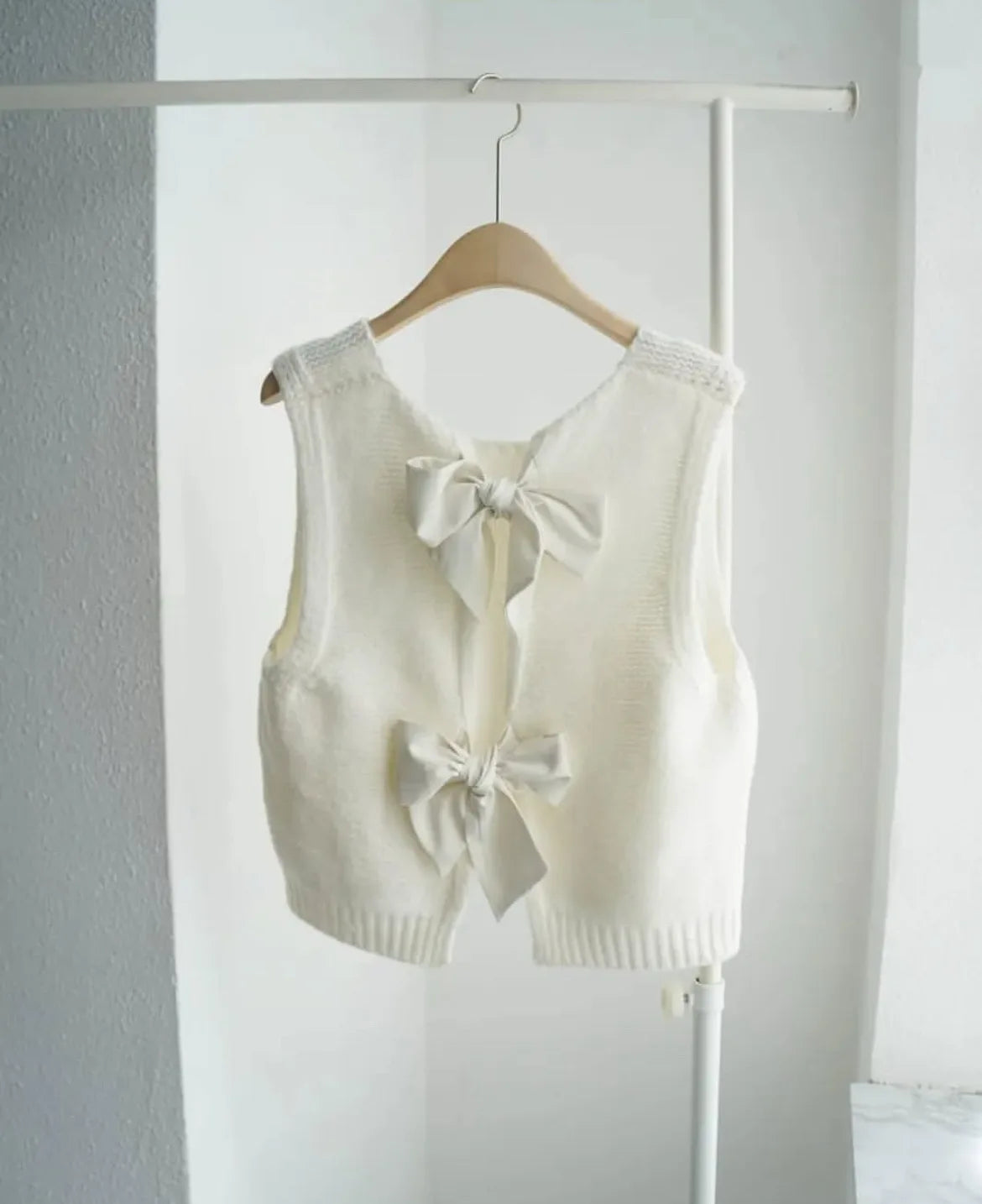 White sleeveless vest with bow details on a hanger against a white wall.