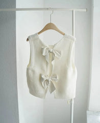 White sleeveless vest with bow details on a hanger against a white wall.