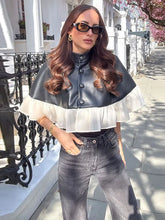 Woman wearing a black leather cape over a white blouse and gray jeans on a sunny street.