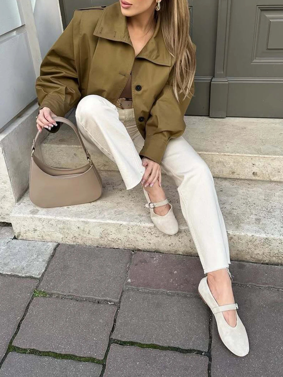 Woman sitting on steps wearing a green jacket, white pants, and beige shoes, holding a beige handbag.