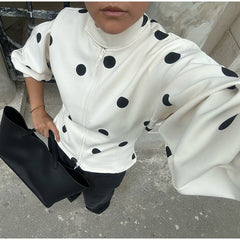 Person wearing a white hoodie with black polka dots, holding a black bag on a concrete surface.