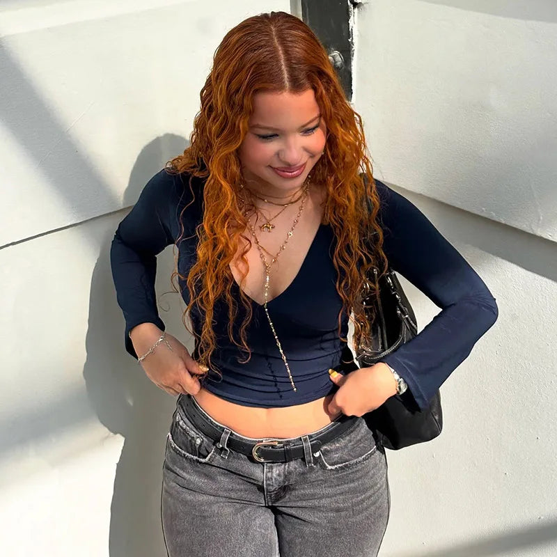 Woman with long red hair wearing a navy blue top and gray jeans against a white wall.