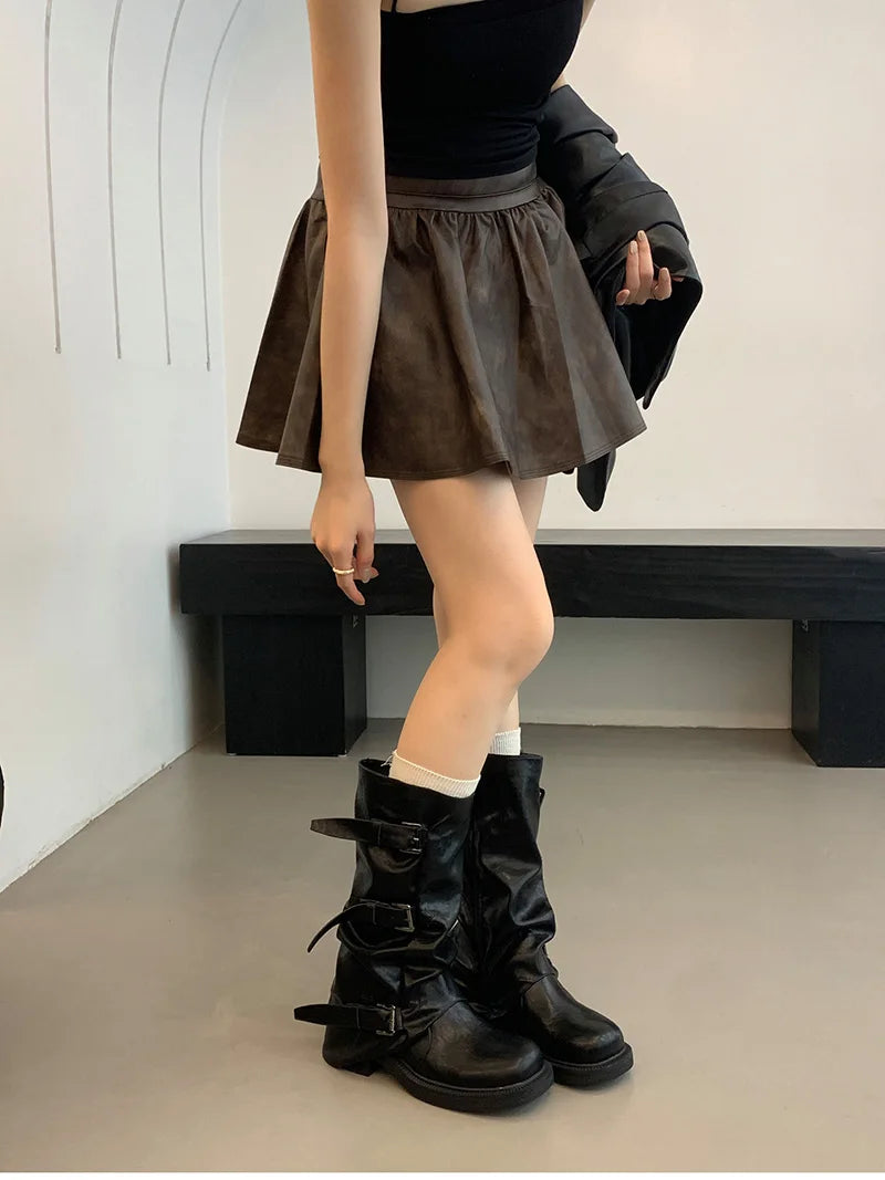 Person wearing a black top, brown skirt, and black boots in a minimalistic setting.