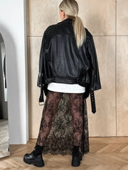 Person wearing a black leather jacket and sheer lace skirt in a room with wooden flooring.