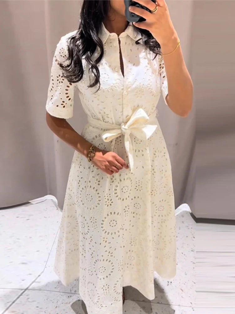 Woman wearing a white lace dress with a bow tie in a minimal indoor setting.