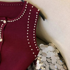 Maroon cardigan with pearl embellishments on a hanger next to silver sequin fabric.