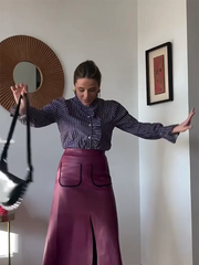 Woman in a striped shirt and purple skirt standing in a room with a mirror and framed picture on the wall.