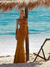 Woman in a mustard yellow dress standing on a beach with turquoise water and a thatched roof in the background.