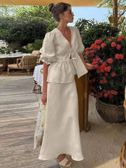 Woman in a white outfit standing outdoors near a table and chairs with a potted plant in the background.