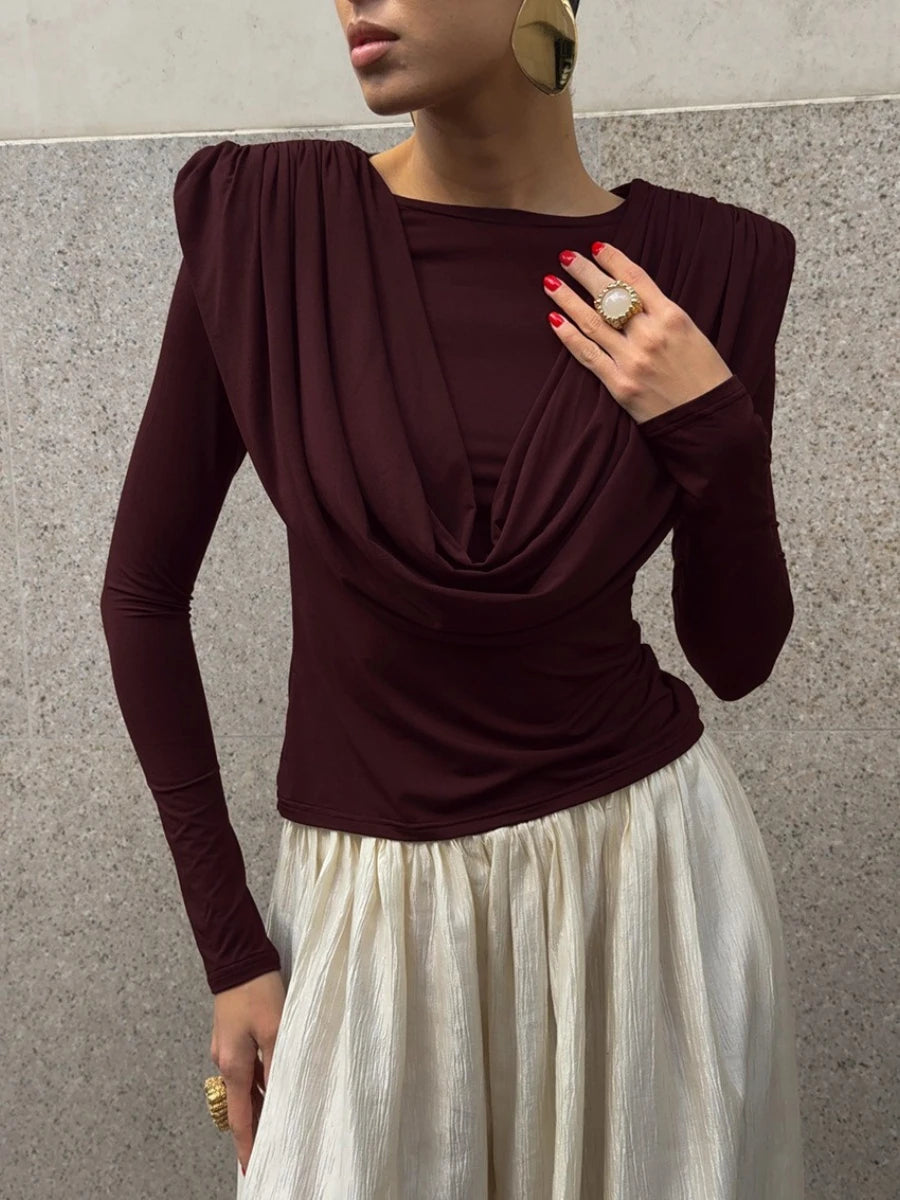 Woman wearing a burgundy top with a draped design and a white skirt against a gray wall.