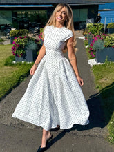 Woman in a white polka dot dress standing outdoors with flowers and water in the background