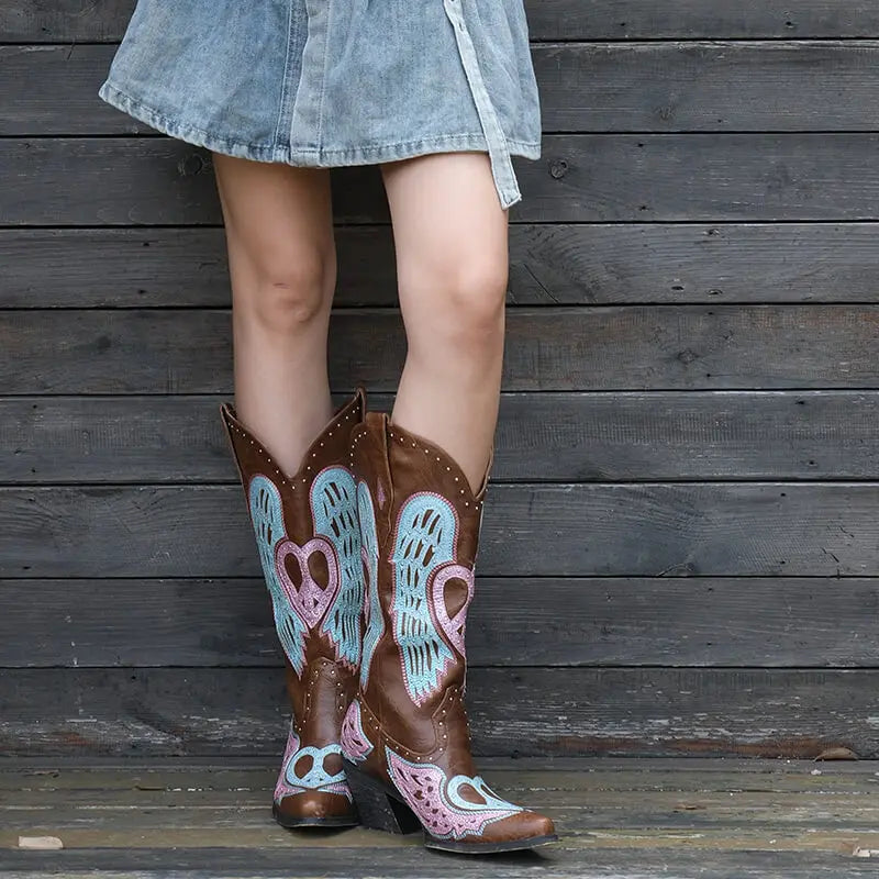 Person wearing decorative cowboy boots with heart and wing designs against a wooden background