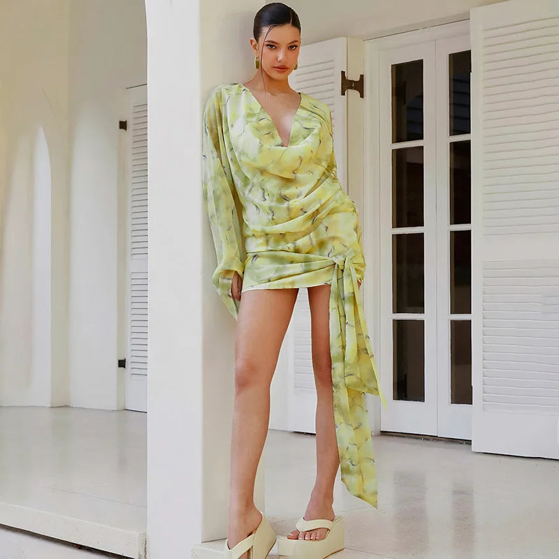 Woman in a green and yellow patterned dress standing in a room with white walls and doors.