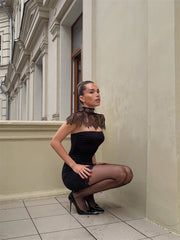 Woman in a black dress with lace details crouching against a building wall.