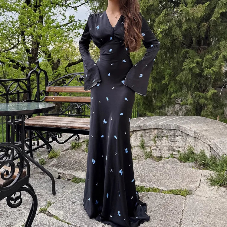 Woman in a black dress with white patterns standing outdoors near a stone wall and bench.