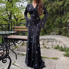 Woman in a black dress with white patterns standing outdoors near a stone wall and bench.