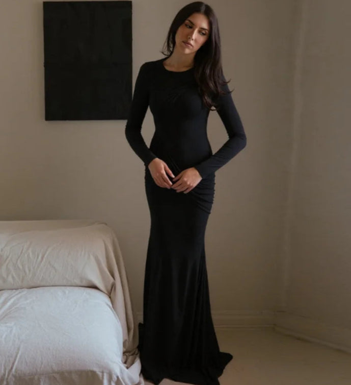 Woman in a black dress standing in a minimalistic room with a bed and a painting on the wall.