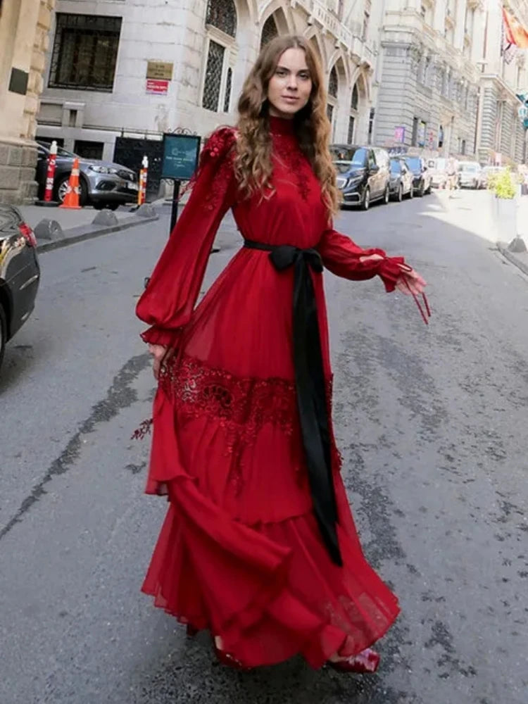 Woman in a red dress with long sleeves and black belt standing on a city street.