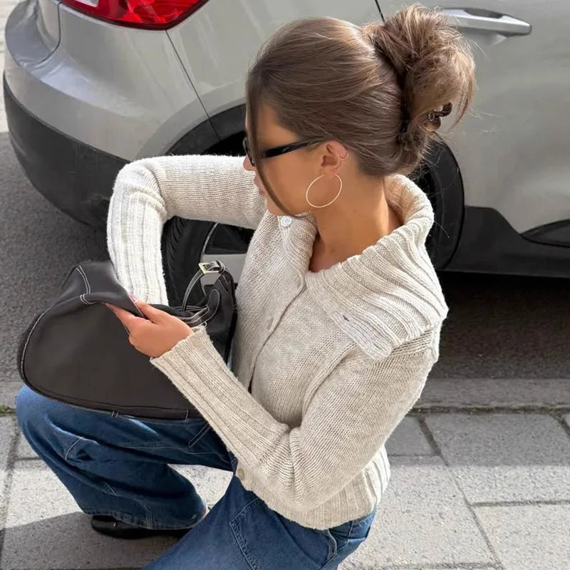 Woman in a beige sweater and blue jeans crouching next to a car, holding a phone.
