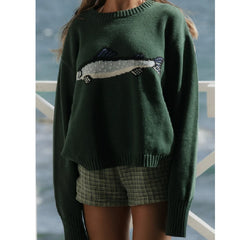 Person wearing a green sweater with a fish design by a body of water.