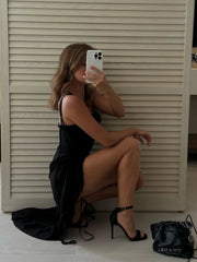 Woman in a sleeveless black party dress with asymmetrical cut taking a mirror selfie against a wooden shutter.