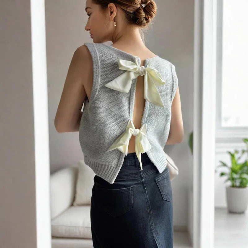 Woman wearing a light gray sleeveless top with a bow detail, standing in a room with a neutral color scheme.