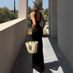Woman in a black backless maxi dress with ankle-length flow and a straw hat holding a woven bag on a modern patio.