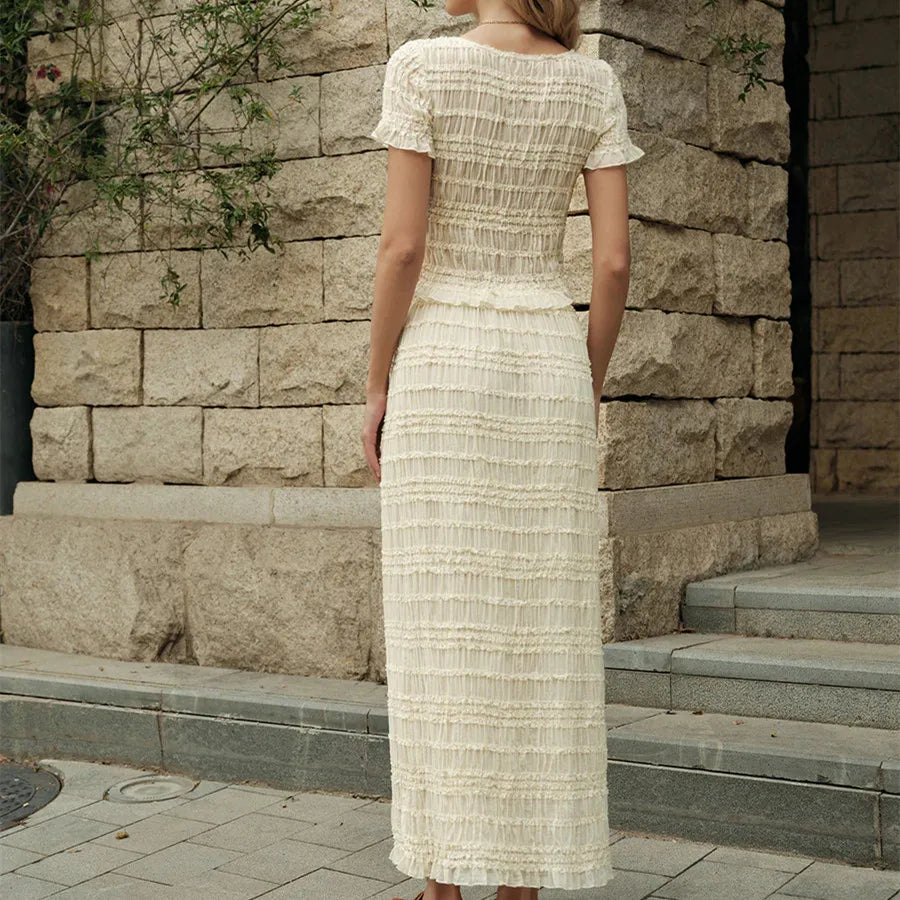Backside view of a woman wearing a cropped top paired with a high-waisted maxi skirt for beachwear, standing in front of a stone wall.